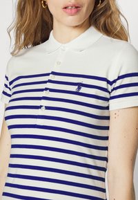 Biała koszulka polo z poziomymi granatowymi paskami, krótkimi rękawami i klasycznym kołnierzykiem. Posiada trzy guziki oraz haftowane logo.
