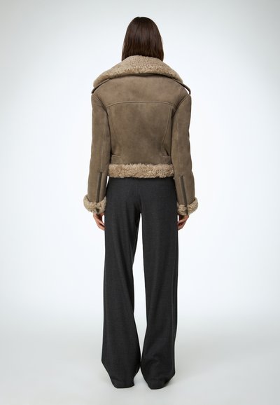 Veste aviateur en cuir marron doublée en peau d'agneau avec un large col et des détails aux poignets, associée à un pantalon large gris foncé. Design minimaliste.