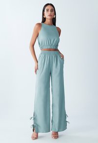 Svetlo teal dvodelni komplet, ki vključuje brezrokavni crop top in široke hlače. Hlače imajo stranske vezi in elastičen pas za udobje.