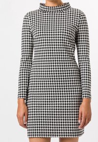 Robe à motifs pied-de-poule noir et blanc, avec des manches longues et un col haut et replié. Fabriquée en tissu extensible pour un look ajusté.