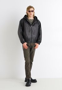 Chaqueta de cuero negra con cremallera y capucha, pantalones verde oliva y zapatos negros gruesos. El modelo lleva unas gafas de sol estrechas y collares en capas.