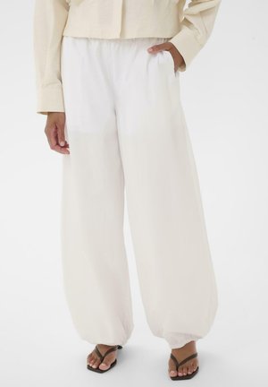 Hvid bukser med vide ben og samlede ankler, båret med en cremefarvet jakke, hånd i lommen og sorte thongsandaler på en ensfarvet baggrund.