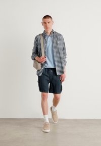 Tenue décontractée comprenant une chemise à carreaux à boutons, un polo bleu, un short marine, des chaussettes blanches et des baskets beiges, avec un sac à bandoulière beige.