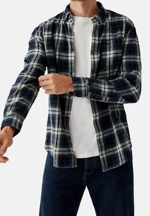 Mann, der die Ärmel eines blau-grau karierten Hemdes mit Knöpfen über einem weißen T-Shirt hochkrempelt, trägt dunkle Jeans, vor einfachem Hintergrund.