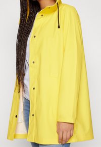 Imperméable jaune en matériau étanche, doté d'une capuche, de boutons-pression et de poches latérales. Texture lisse avec une coupe décontractée.