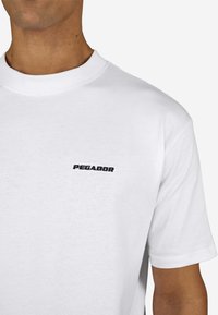 Weißes kurzärmeliges T-Shirt aus Baumwolle, mit einem kleinen schwarzen "PEGADOR"-Logo auf der linken Brustseite und einem Rundhalsausschnitt.