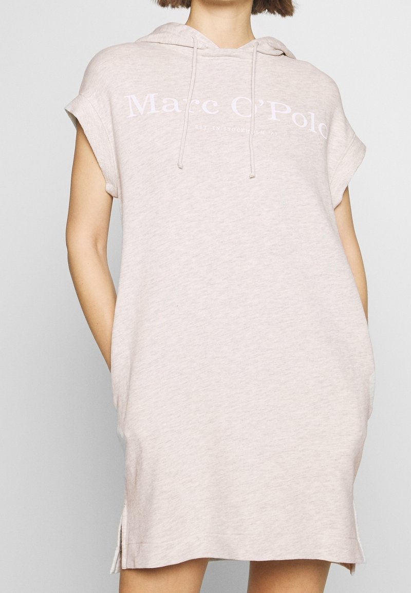 Femme portant une robe à capuche sans manches beige clair avec le texte "Marc O’Polo" sur la poitrine et les mains dans les poches latérales.