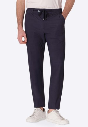 Uomo che indossa pantaloni slim fit blu scuro con coulisse e bottone, sneakers bianche e una camicia chiara infilata.