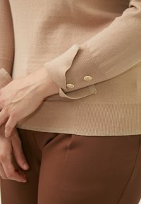 Beige trui met ribbeltextuur en gouden accentknopen op de manchetten, gecombineerd met bruine broek. Heeft een getailleerd ontwerp en zachte stof.