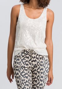 Débardeur blanc pailleté avec un décolleté arrondi, associé à un pantalon beige et noir à motifs géométriques.
