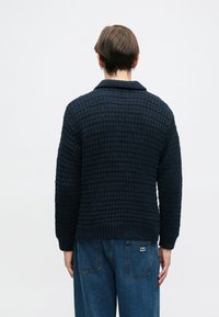 Sötét tengerészkék kötött pulóver texturált mintával, galléros nyakkal és bordázott mandzsettákkal. Kék farmernadrággal viselve.