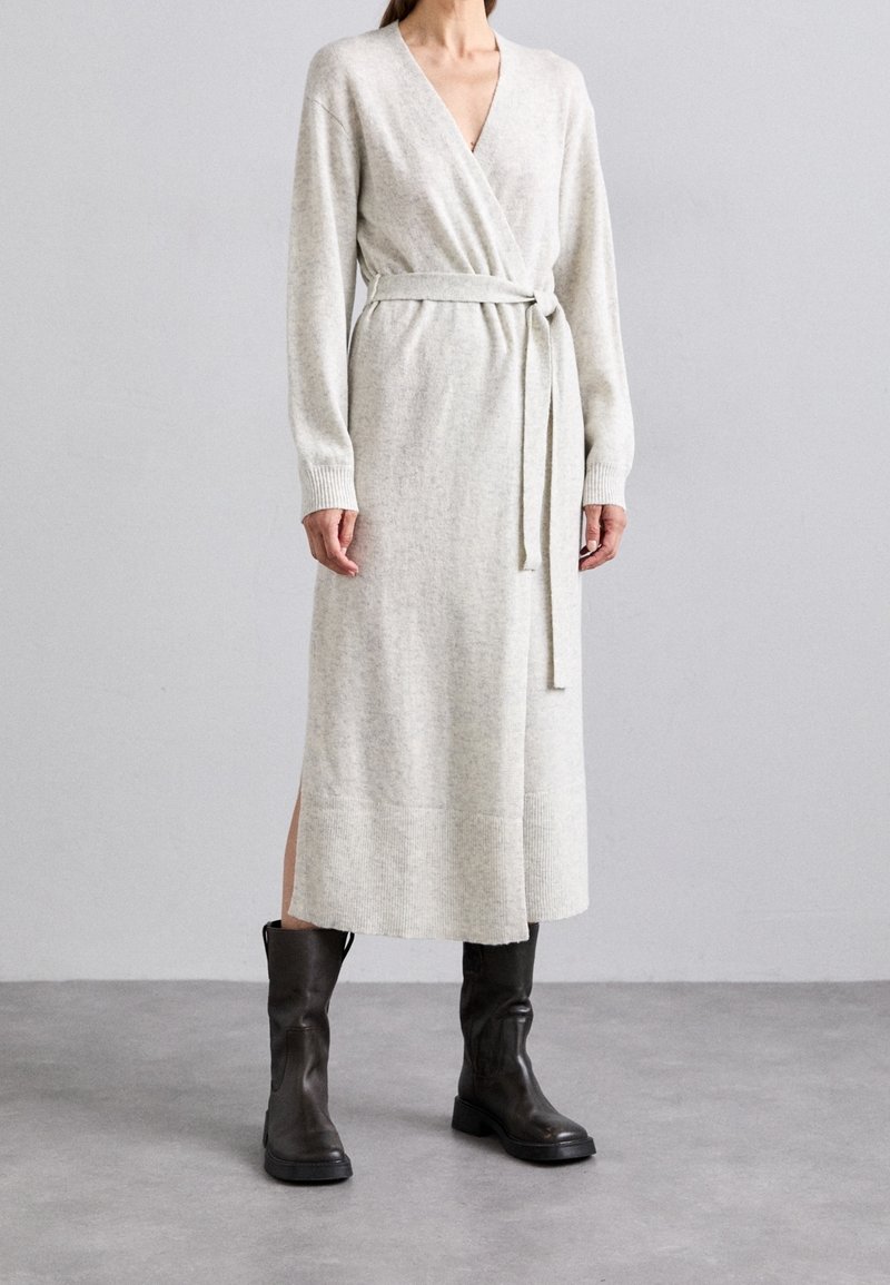 Femme porte une longue robe en maille gris clair ceinturée et des bottes en cuir noires mi-mollet, debout sur un sol gris devant un mur uni.