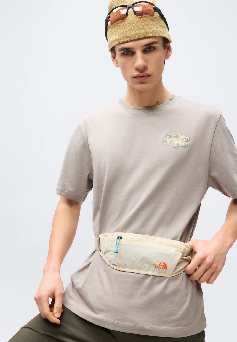Young man wearing beige beanie with sunglasses, gray t-shirt with van logo, and beige North Face waist bag adjusting the pouch strap.