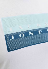 Fehér pamut póló, amelyen egy grafikával díszített három vízszintes sáv világoskék és sötétkék színekben, kiemelve a "Jack & Jones" márkanevet.