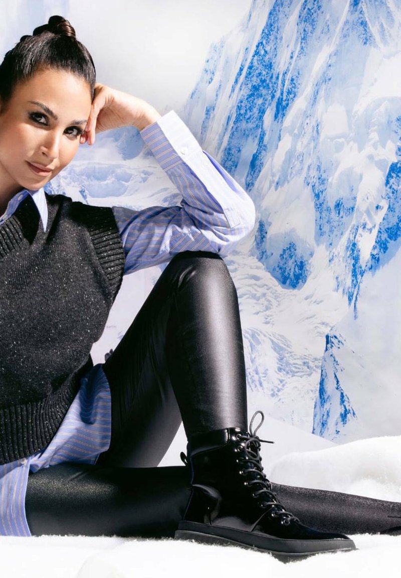 Kobieta z ciemnymi włosami w kok z czarnym bezrękawnikem na niebieskiej w paski koszuli, czarnych legginsach i czarnych sznurowanych butach, siedząca na tle śnieżnych gór.