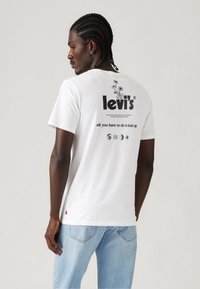Weißes Baumwoll-T-Shirt mit bedrucktem schwarzen Text und Artwork auf der Rückseite, mit lockerer Passform und kurzen Ärmeln. Kombiniert mit hellblauen Jeans.
