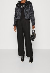 Black quilted puffer jacket, high collar, zip closure, worn over a black top and wide black trousers. Holding a small textured black handbag.