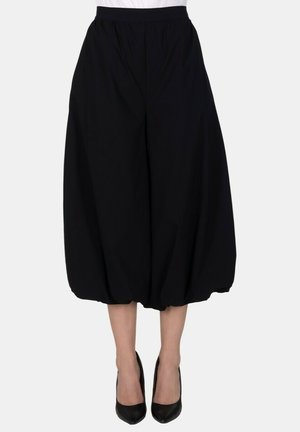 Pantaloni ballon neri a gamba larga indossati con scarpe nere con tacco alto, tagliati sopra le caviglie, su uno sfondo semplice.