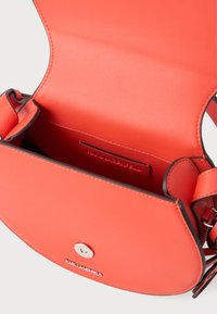 Oranssi nahkainen olkalaukku, jossa on pyöreä läppä, hopeiset yksityiskohdat ja etupuolella kohokuvioitu logo. Sisätilat koostuvat yhdestä osastosta.