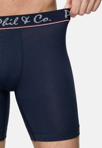 Marinblå boxerkalsonger i stretchmaterial med en mjuk midjeband med logotyp och röda/vita accenter. Slät textur, längd till mitten av låret.