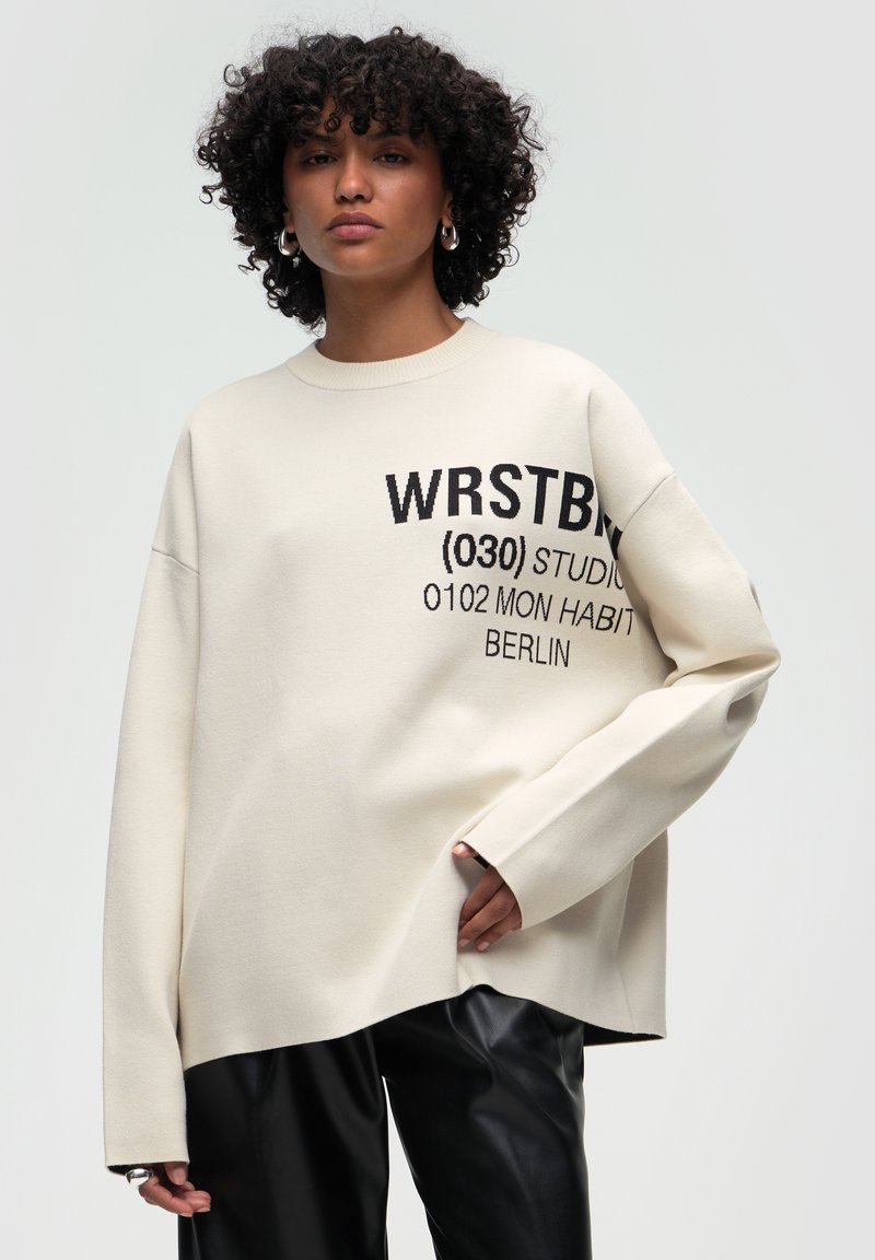 Krämfärgad sweatshirt med svart grafisk text, som har en avslappnad passform, sänkta axlar och ribbad halsringning. Matchas med svarta byxor.