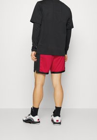 Persona con una camiseta de manga larga negra, pantalones cortos deportivos negros y rojos, calcetines negros y zapatillas blancas de pie frente a un fondo liso.