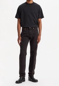 Camiseta negra de manga corta con logotipo en relieve, combinada con jeans rectos de color gris oscuro. El modelo lleva botas de cuero negras hasta el tobillo.
