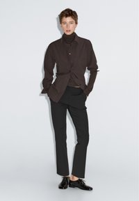 Zwarte button-up shirt over een bruine coltrui, gecombineerd met zwarte tapered broek en glanzende zwarte schoenen, tegen een effen lichte achtergrond.