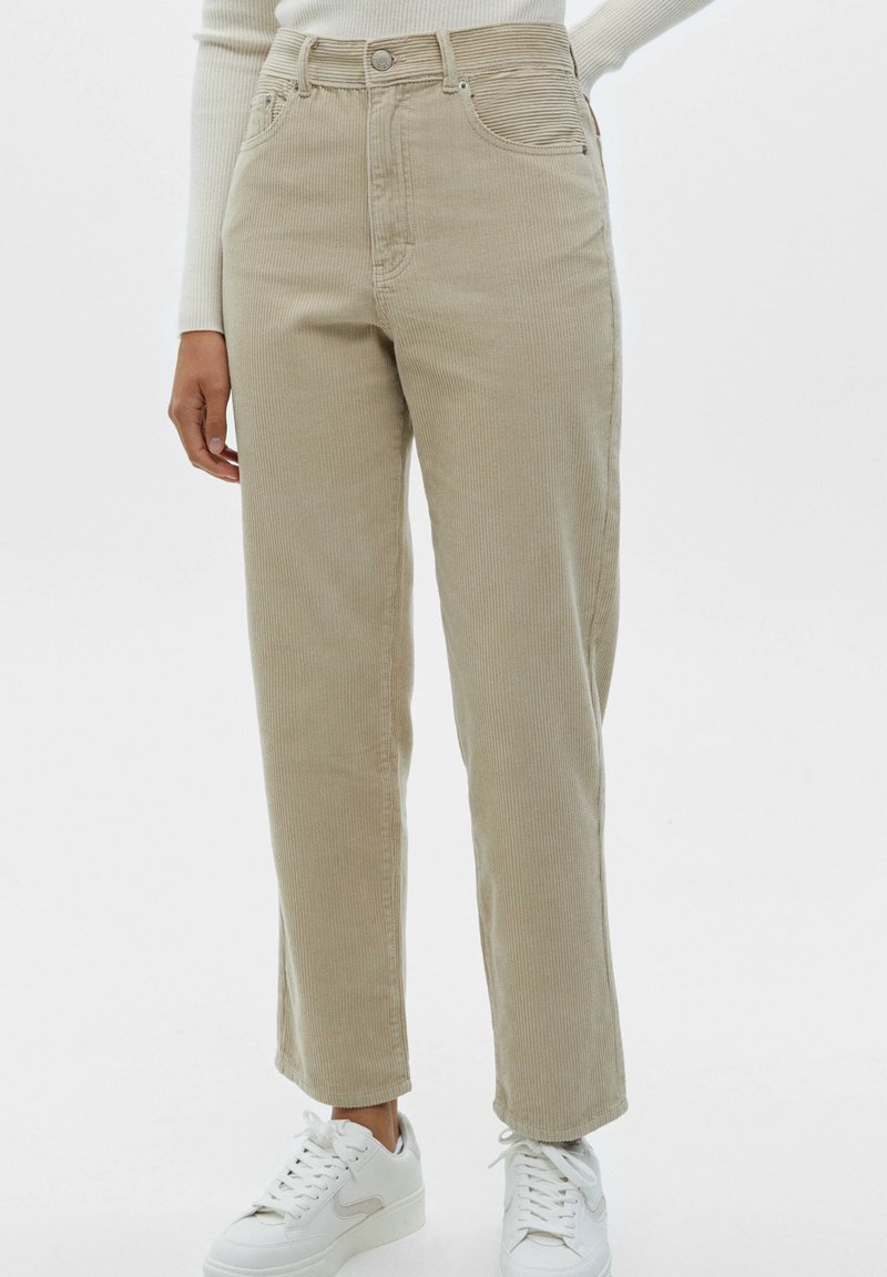Pantalon en velours côtelé beige à jambes droites, porté avec un haut blanc à manches longues côtelé et des baskets blanches sur fond blanc.
