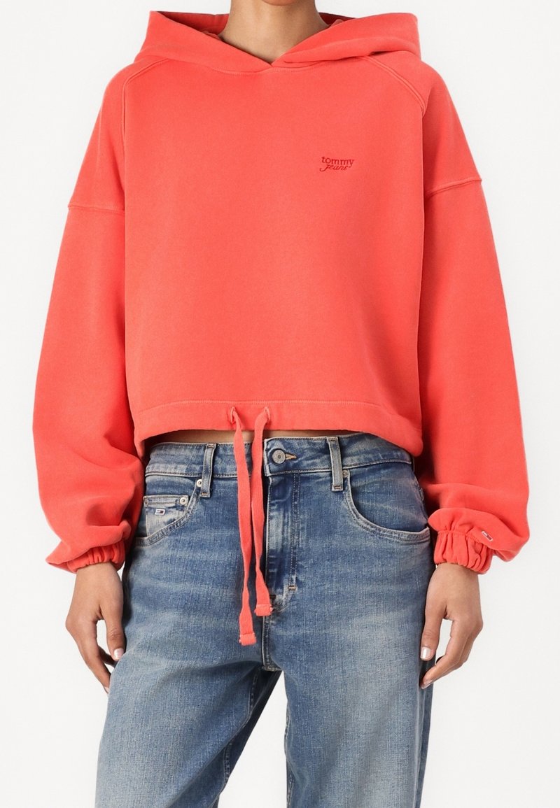 Sudadera coral recortada con capucha, puños elásticos y dobladillo con cordón. Cuenta con un pequeño logo en el pecho y una textura suave de algodón.