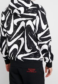 Hoodie med ett svart och vitt virvlande abstrakt mönster. Har ribbad mudd och nederkant, samt röd logotyp på nedre ryggen.