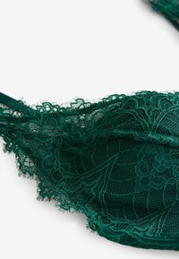 Tamnozelena čipkasta brusnica s složenim cvjetnim uzorcima, prozirnom tkaninom i nježnim obrubom. Ima podešive naramenice i mekanu teksturu.