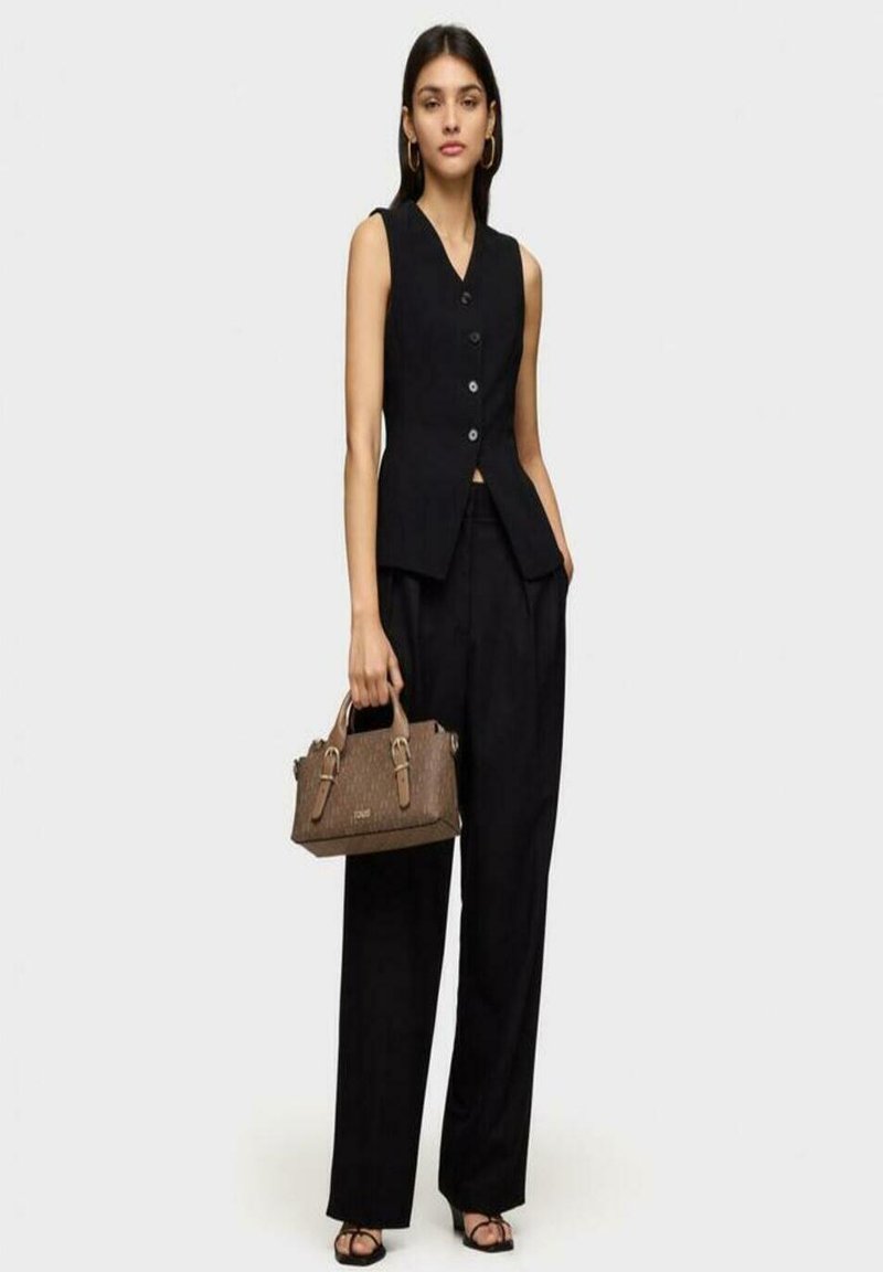 Chaleco negro sin mangas con botones, combinado con pantalones anchos negros. Un bolso de cuero texturizado marrón con correas completa el look.