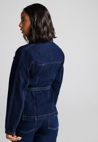 Mörkblå denimjacka med en figursydd design, synliga sömnadsdetaljer och midjebälte. Bakdelen har en skuldra och sidfickor.