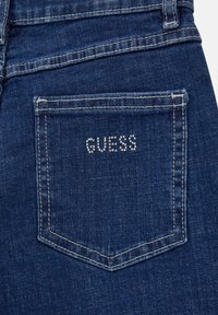 Donkerblauwe denim jeans met een achterzak waarin het woord "GUESS" is versierd met heldere strassstenen, vergezeld van gele stiksels.
