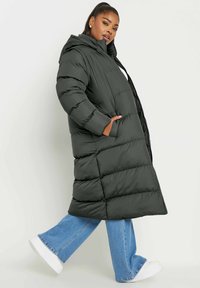 Abrigo largo acolchado de color verde oscuro con capucha, que cuenta con un cierre frontal, bolsillos laterales y una silueta cuadrada, combinado con unos pantalones vaqueros de pierna ancha de color azul claro.