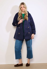 Blonde Frau trägt einen marineblauen zweireihigen Mantel, grünes Oberteil, blaue Cropped-Jeans und braune Loafers und steht vor einem schlichten Hintergrund.