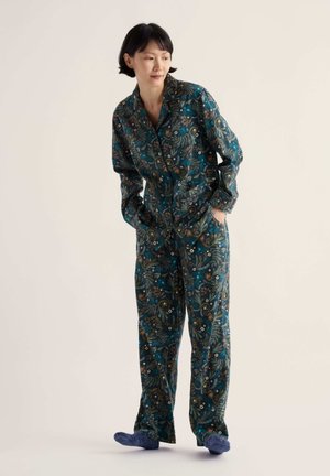Femme en pyjama à manches longues vert foncé à motif floral, debout, les mains dans les poches, portant des chaussettes bleues sur fond uni.