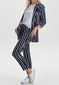 Femme portant un costume à rayures verticales noires et blanches avec un chemisier blanc texturé et des baskets blanches, debout avec une jambe croisée.