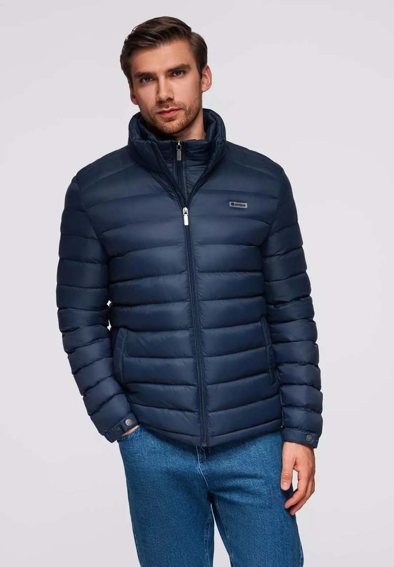 Marineblaue Steppjacke mit horizontalem Steppmuster, hohem Kragen und Reißverschluss. Verfügt über ein kleines Logo und eine glatte, glänzende Textur.