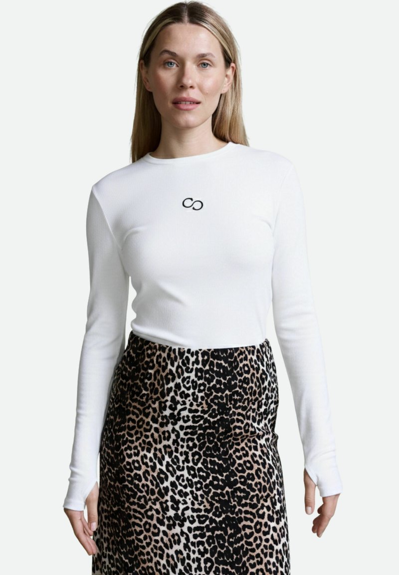 Weißes Langarmoberteil mit gerippter Textur und schwarzem Unendlichkeitslogo auf der Brust, kombiniert mit einem Rock im braunen und schwarzen Leopardenmuster.