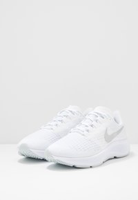 Scarpe da ginnastica bianche con una tomaia in rete traspirante, superficie testurizzata e uno swoosh Nike riflettente color argento sul lato. Suola in gomma.