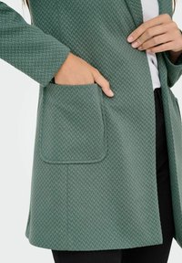 Manteau vert texturé avec un motif en chevrons, avec une poche sur le côté gauche et une coupe droite. Conçu pour le confort et la facilité.