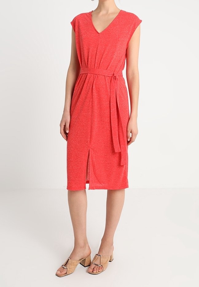 Robe rouge jusqu'aux genoux avec un col en V, des manches courtes en forme de petites ailes et une ceinture à nouer. Comprend une fente à l'avant et un tissu texturé. Portée avec des sandales beiges.