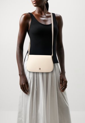 Femme portant un haut noir sans manches et une jupe plissée blanche, tenant un sac bandoulière crème avec un logo doré à l'avant.