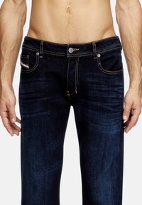 Jeans de mezclilla oscura con un ajuste slim, adornados con costuras naranjas, un botón plateado y bolsillos. La tela tiene una textura suave.