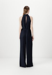 Navy blue jumpsuit, pleated fabric, open back with a keyhole detail, wide legs, and a fitted waist. Minimalist design with textured finish.