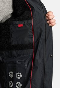 Musta takki, jossa on pehmeä sisävuori, punainen somiste, useita taskuja ja pyöreitä kangashuoltosymboleja sisätaskussa.