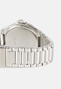 Rustfritt stål klokke med en polert, glatt kasse, sammenkoblet armbånd, og en rund urskive med en teksturert krone. Baksiden viser graverte detaljer.