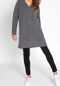 Žena oblečená v šedém žebrovaném dlouhém svetru, černých skinny kalhotách a bílých slip-on teniskách, stojící s jednou nohou vystrčenou dopředu.